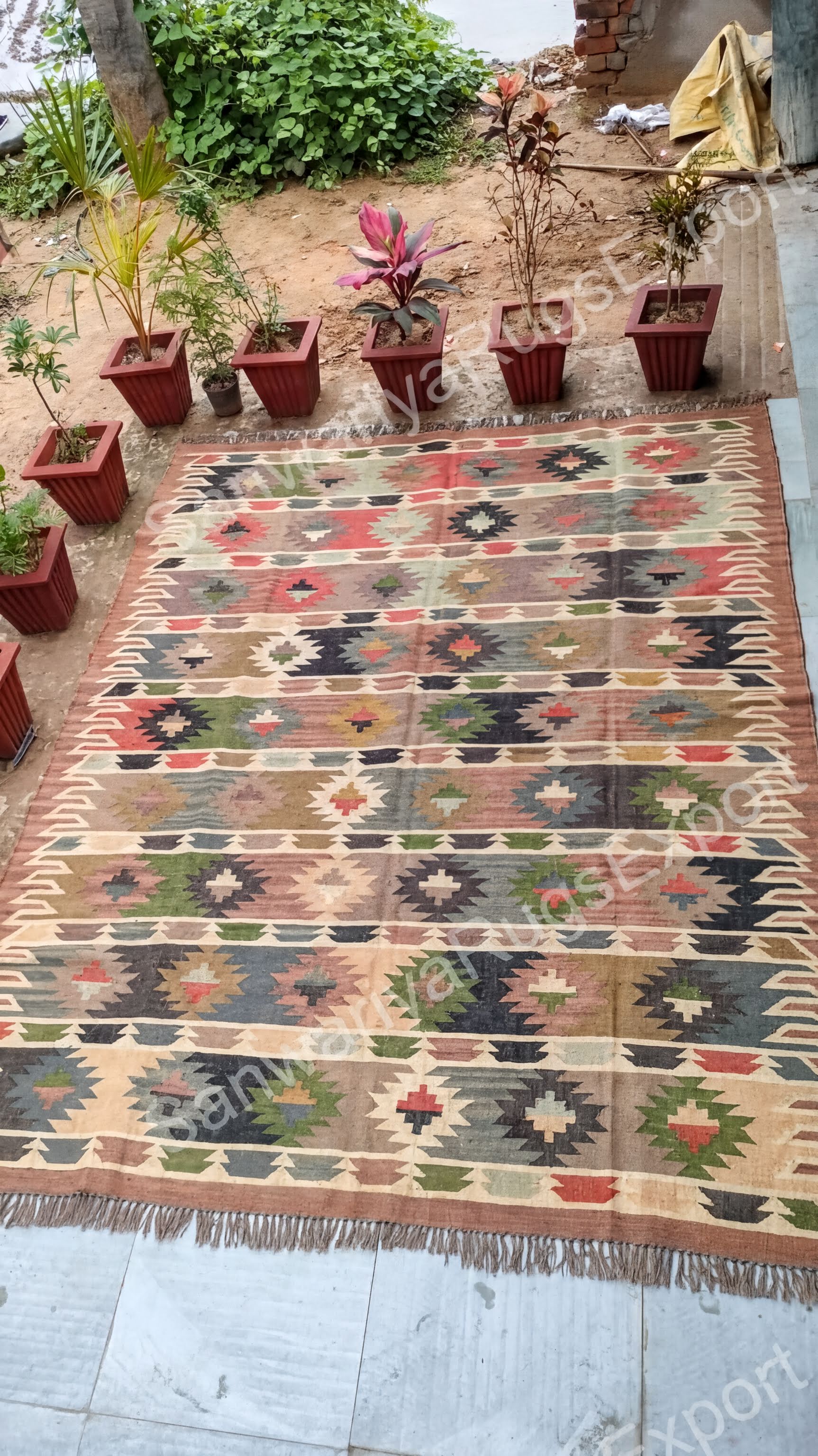 a colorful, patterned rug with a geometric design, surrounded by several potted plants arranged in a garden setting.