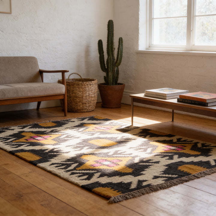 a cozy living room with a beige sofa, a wooden coffee table, and a colorful patterned rug on a wooden floor. A potted cactus plant is placed next to the sofa, and a basket is located nearby. A window allows natural light to illuminate the space, creating a warm and inviting atmosphere.