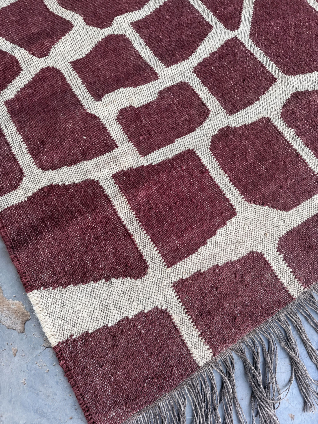 a close-up view of a rug with a geometric pattern in shades of red and white. The rug has a fringed edge and appears to be made of a woven material.