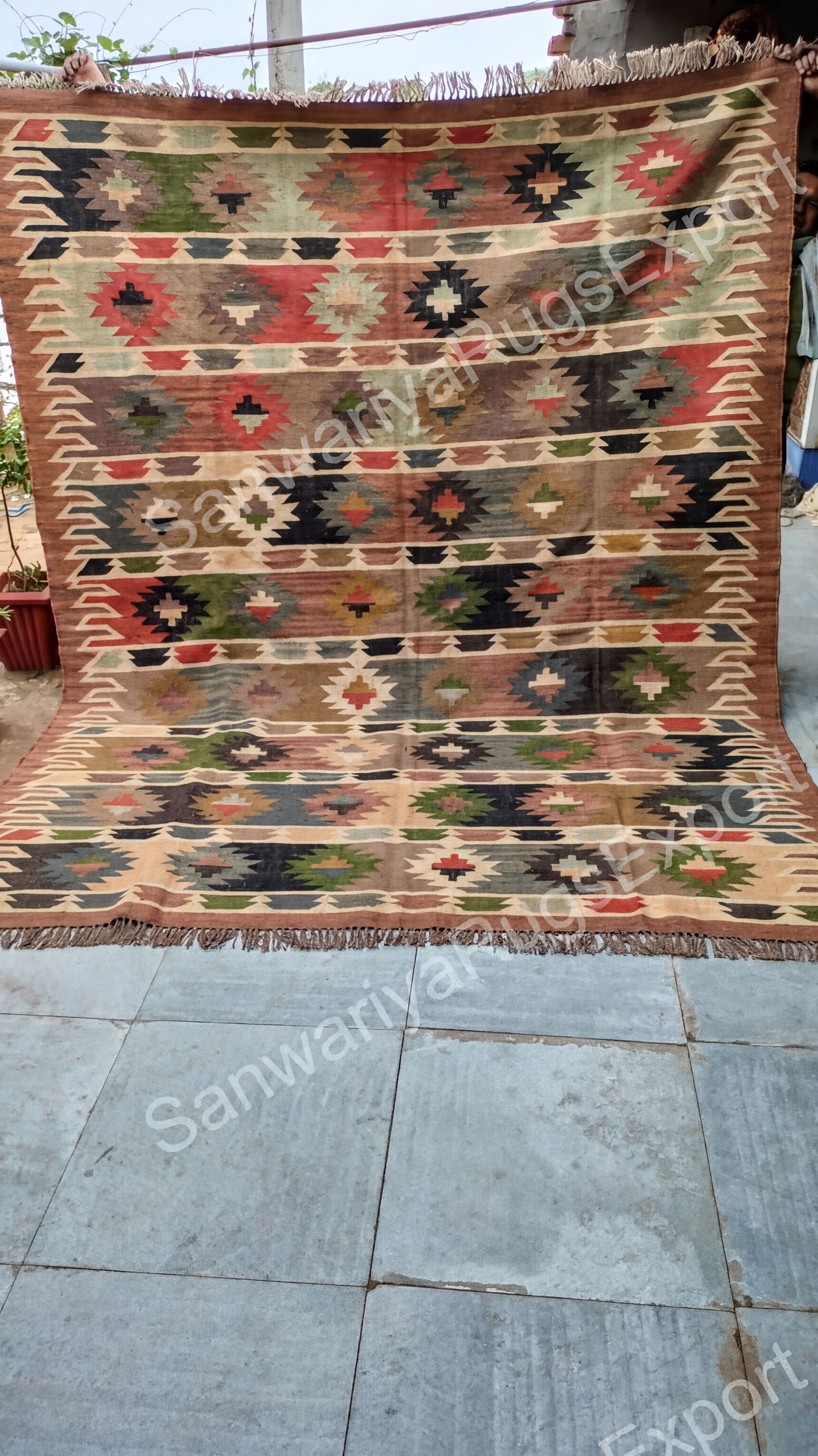 a large, colorful, and intricately patterned rug with a geometric design. The rug is placed on a tiled floor, and there is a potted plant visible in the background.