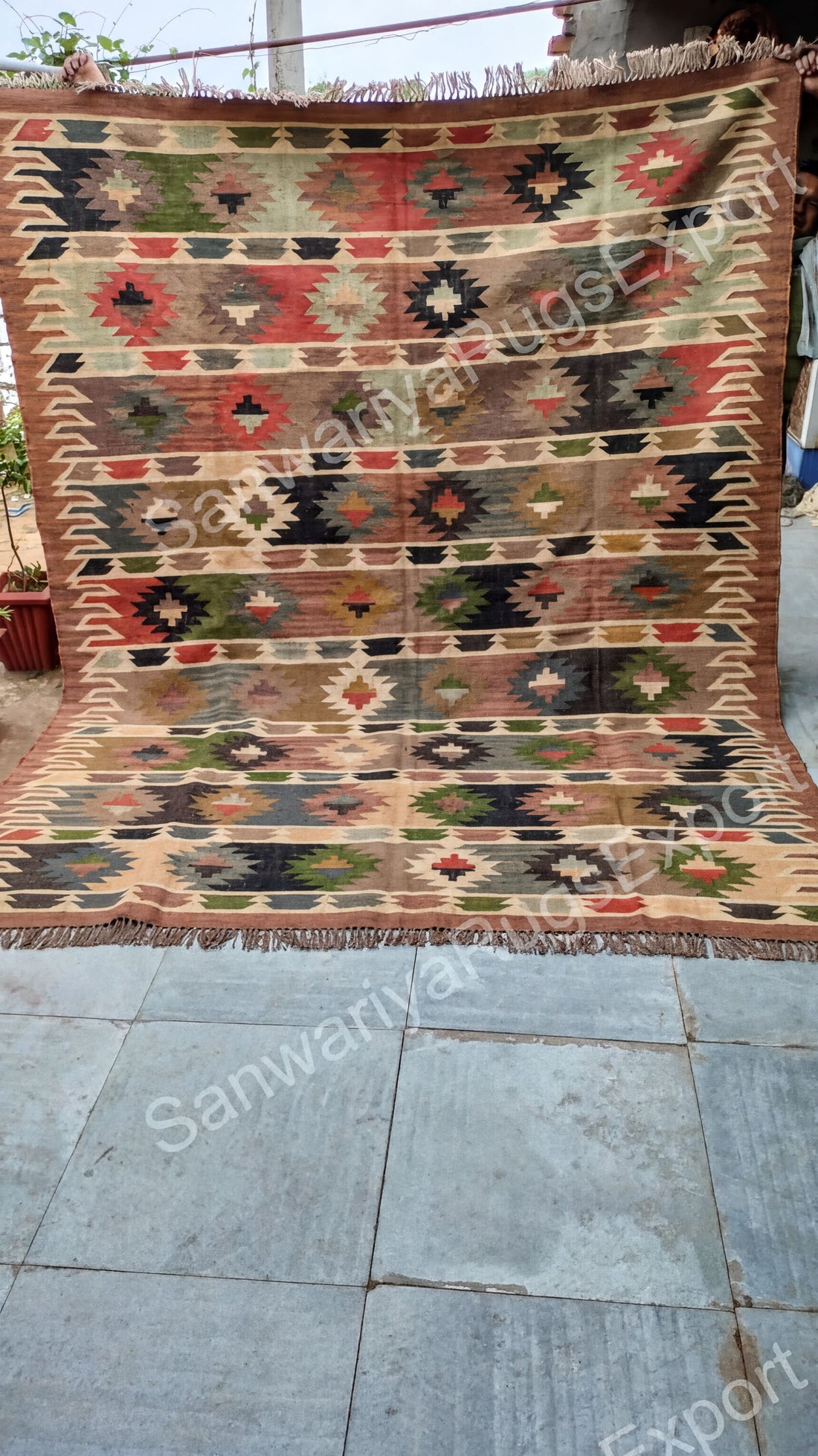 a large, colorful, and intricately patterned rug with a geometric design. The rug is placed on a tiled floor, and there is a potted plant visible in the background.