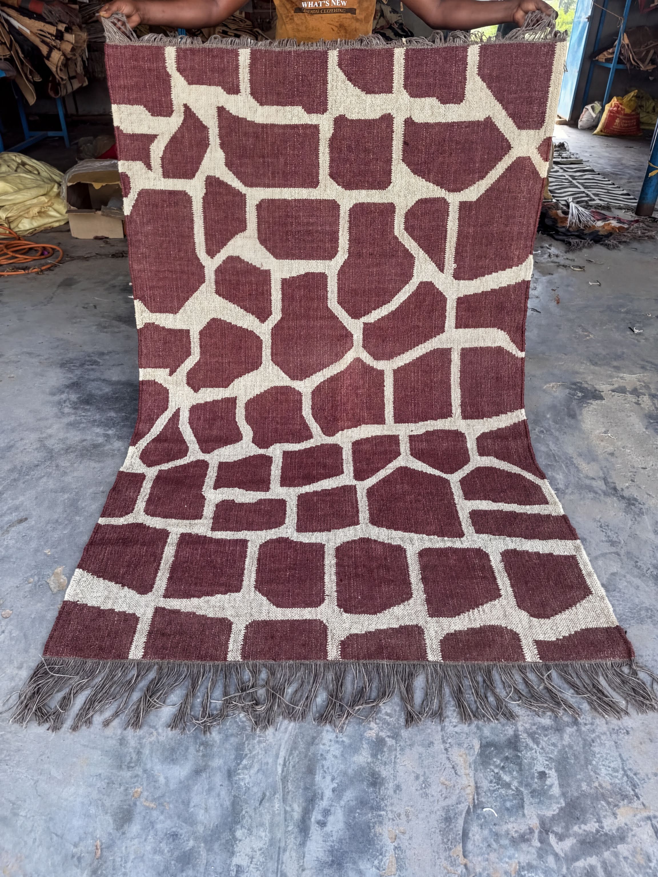 a large, maroon and white patterned rug with a giraffe print design. The rug is placed on a concrete floor, and there are some objects in the background, including a handbag and a suitcase.