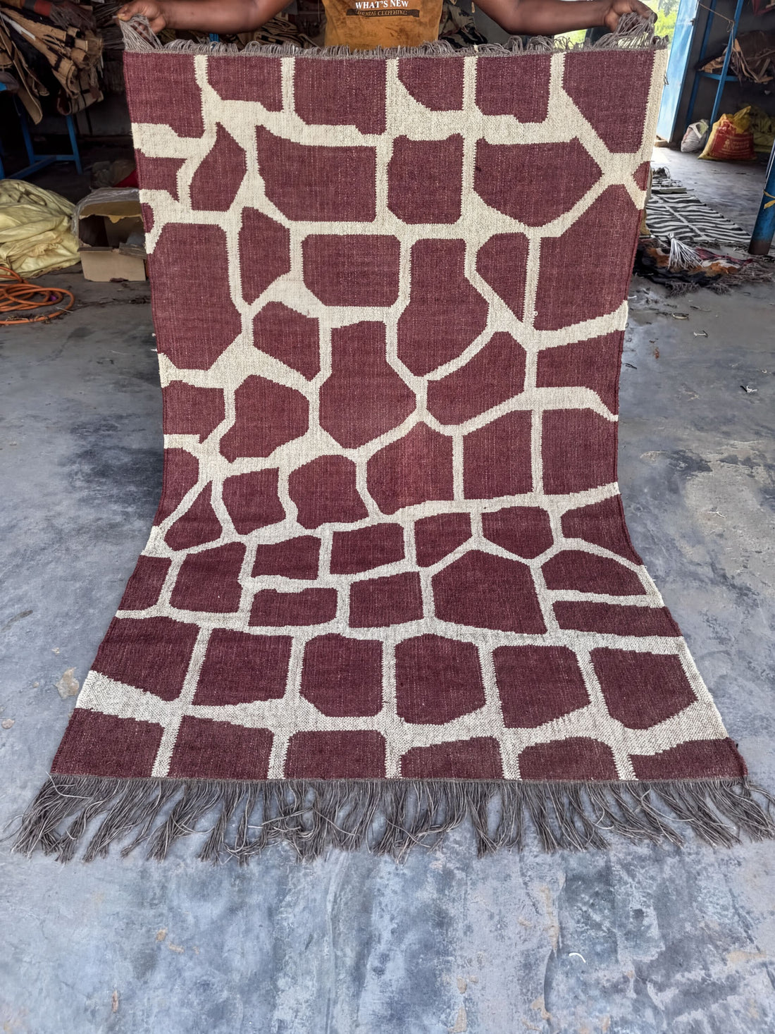 a large, maroon and white patterned rug with a giraffe print design. The rug is placed on a concrete floor, and there are some objects in the background, including a handbag and a suitcase.