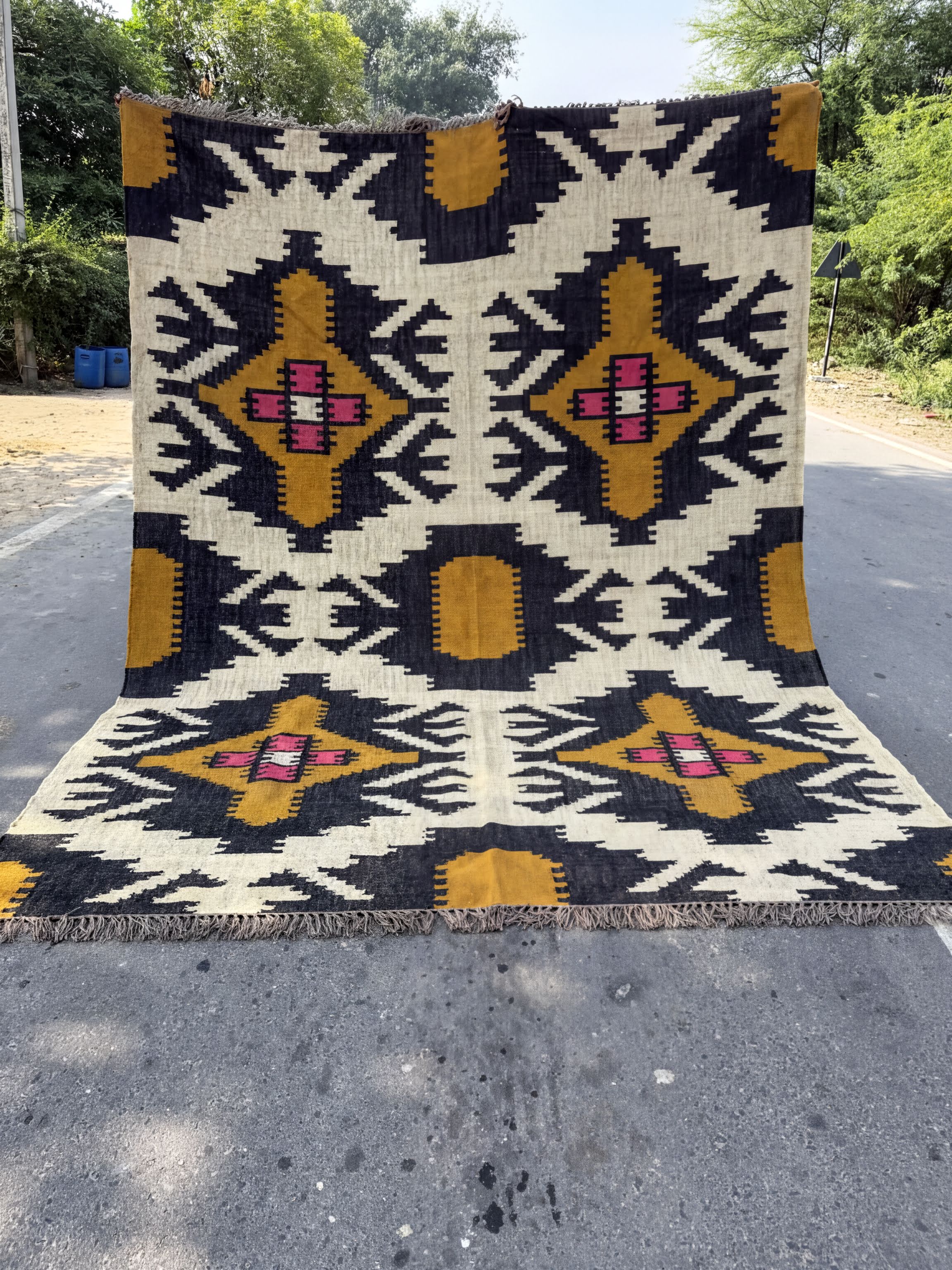 a large, colorful rug with a geometric pattern in shades of black, yellow, and pink. The rug is displayed on a paved surface, possibly a street or sidewalk, with trees and a building visible in the background.