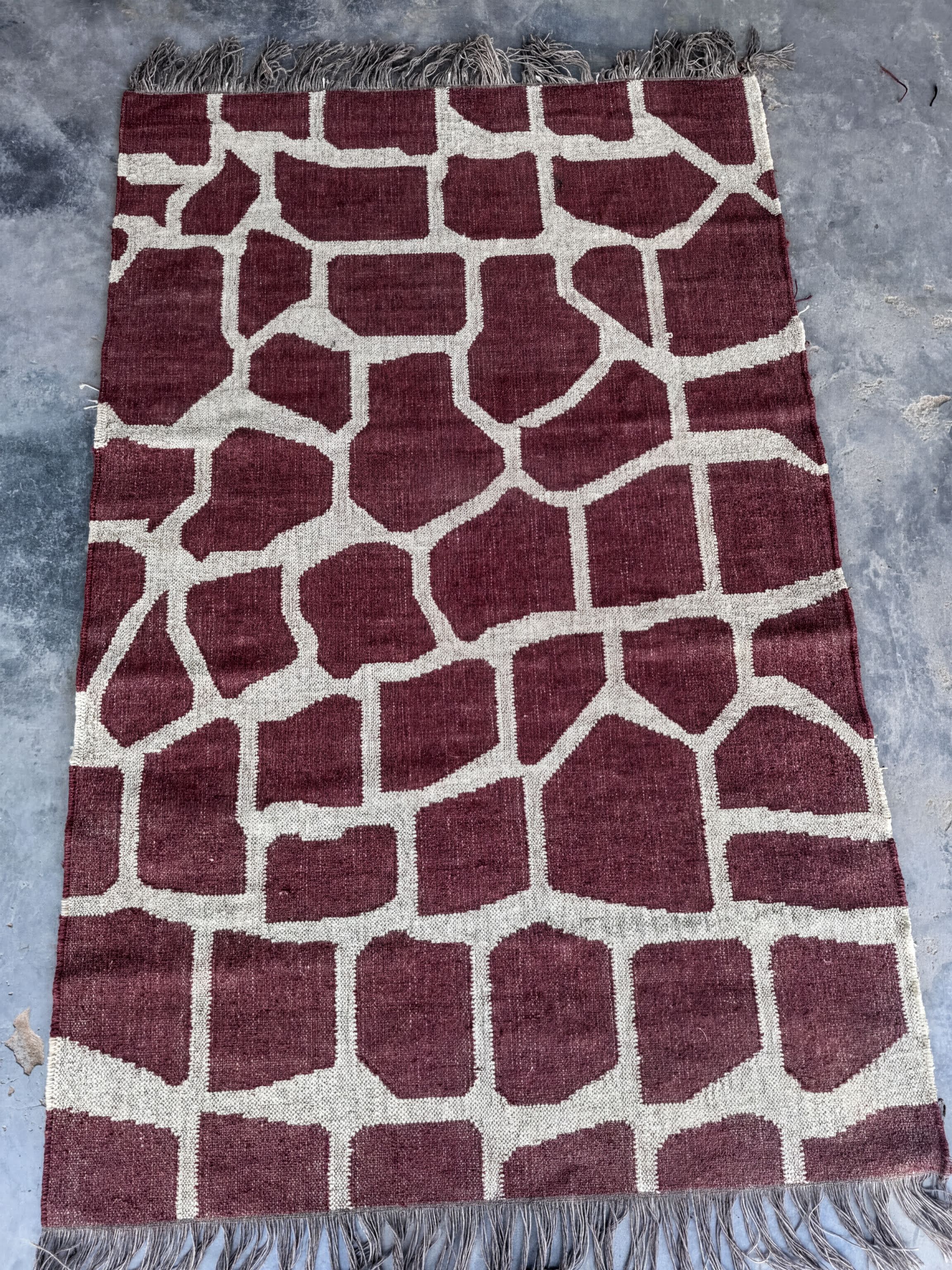 a large, rectangular rug with a unique giraffe print design. The rug has a maroon and white color scheme, with the giraffe pattern composed of various shades of brown and white blocks. The rug is placed on a gray surface, and there is a fringe at the bottom edge.