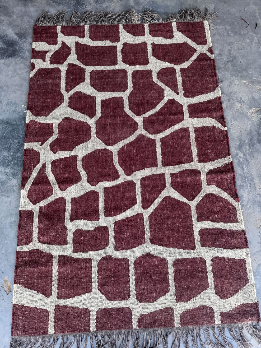 a large, rectangular rug with a unique giraffe print design. The rug has a maroon and white color scheme, with the giraffe pattern composed of various shades of brown and white blocks. The rug is placed on a gray surface, and there is a fringe at the bottom edge.