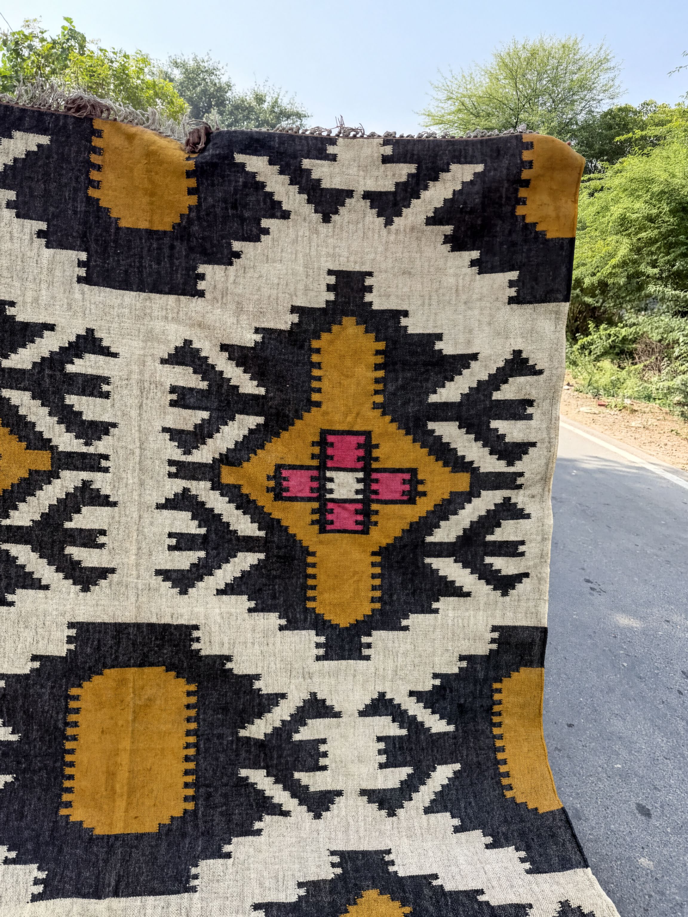a large, colorful rug with a geometric pattern in black, yellow, and pink. The rug is displayed outdoors, with a road and trees visible in the background.