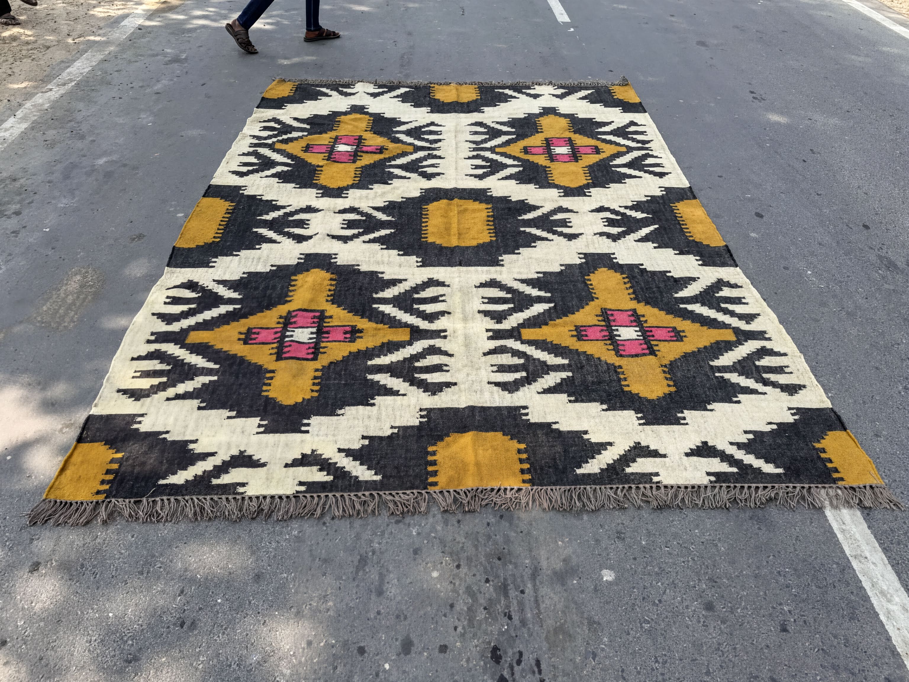 A large, colorful rug with a geometric pattern in shades of yellow, black, and white is spread out on the ground, with a person walking by in the background.