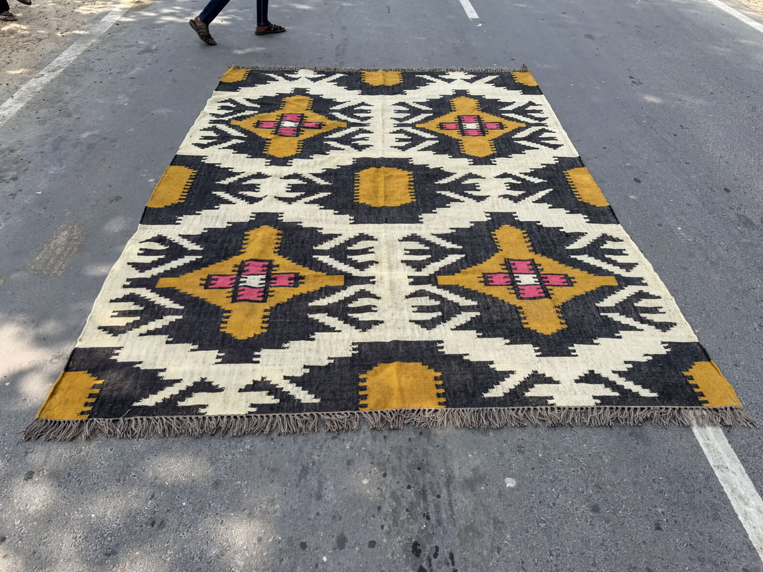 A large, colorful rug with a geometric pattern in shades of yellow, black, and white is spread out on the ground, with a person walking by in the background.