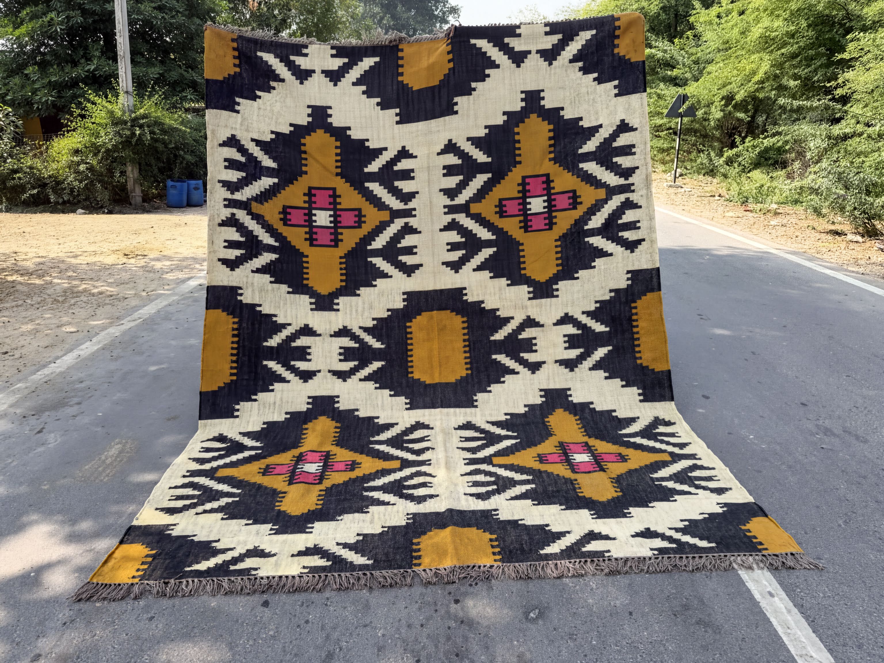 A large, colorful rug with a geometric pattern is displayed on the side of a road, surrounded by trees and bushes.
