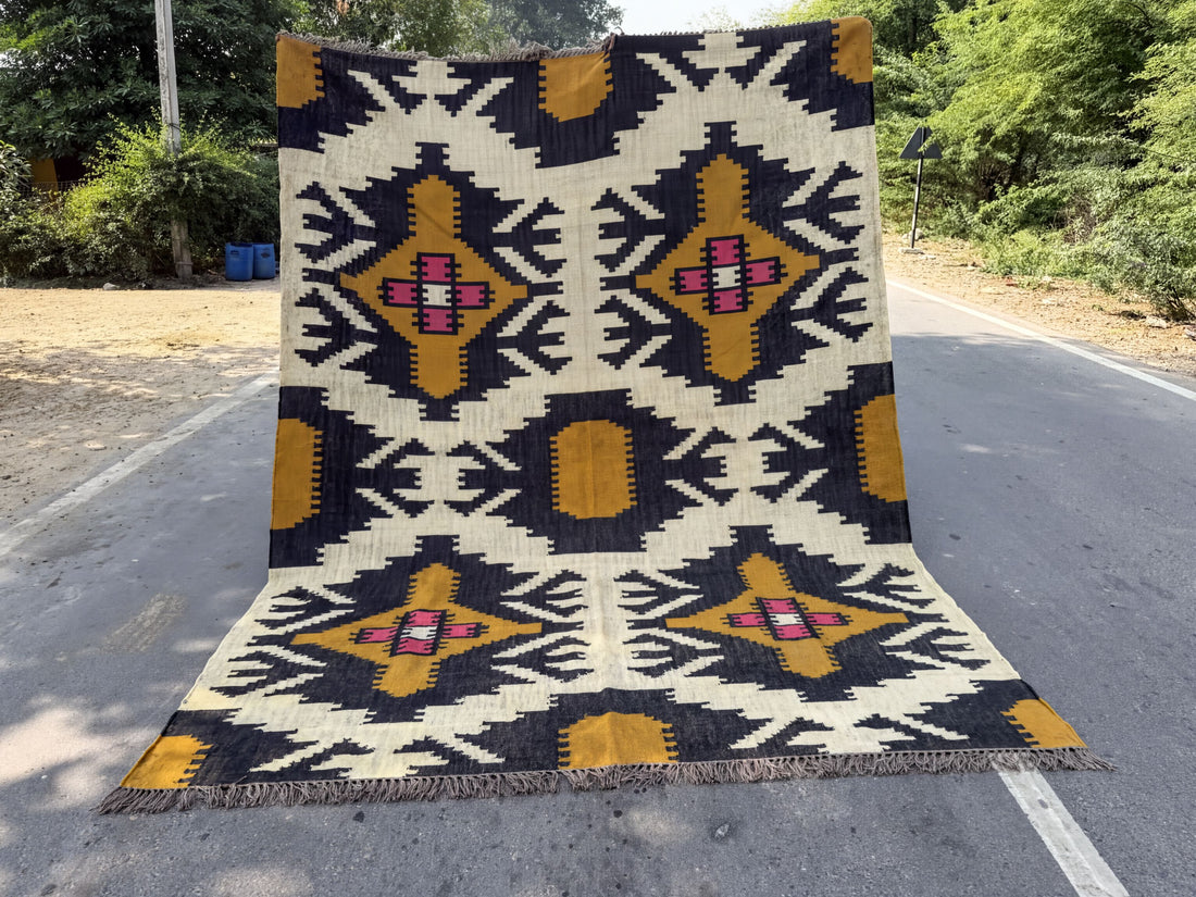 A large, colorful rug with a geometric pattern is displayed on the side of a road, surrounded by trees and bushes.