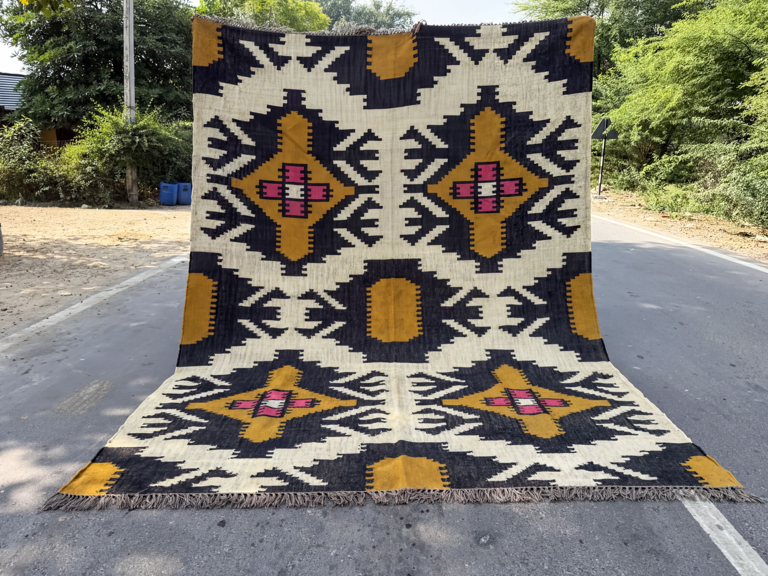 A large, colorful rug with a geometric pattern is displayed on the side of a road, surrounded by greenery.