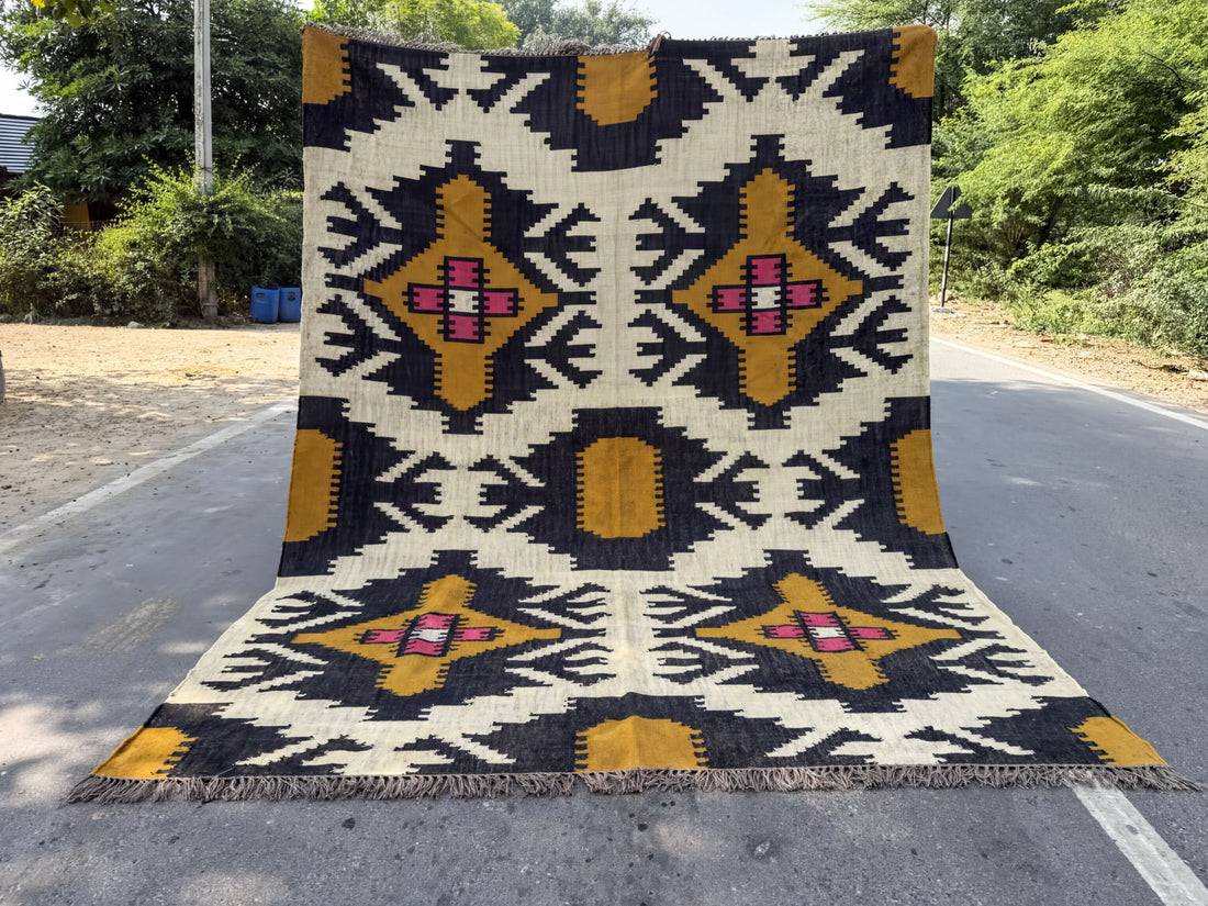A large, colorful rug with a geometric pattern is displayed on the side of a road, surrounded by greenery.